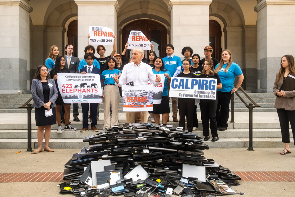 Asm. Bennett at podium, speaking, with pile of e-waste in foreground and supporters in background