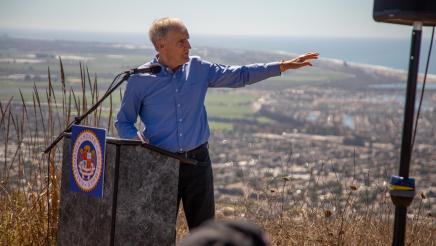 Asm. Bennett at podium, turned to one side and reaching one arm out to land behind him