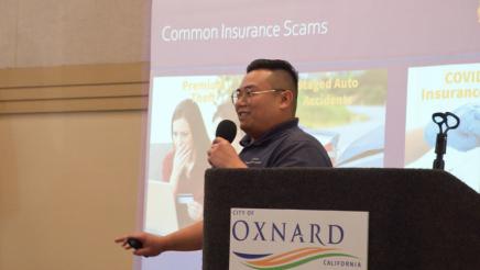 Guest speaker holding microphone, speaking, with large screen in background