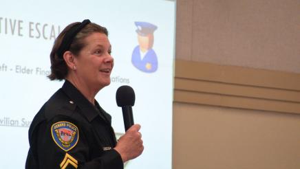 Guest speaker holding microphone, speaking, with large screen in background